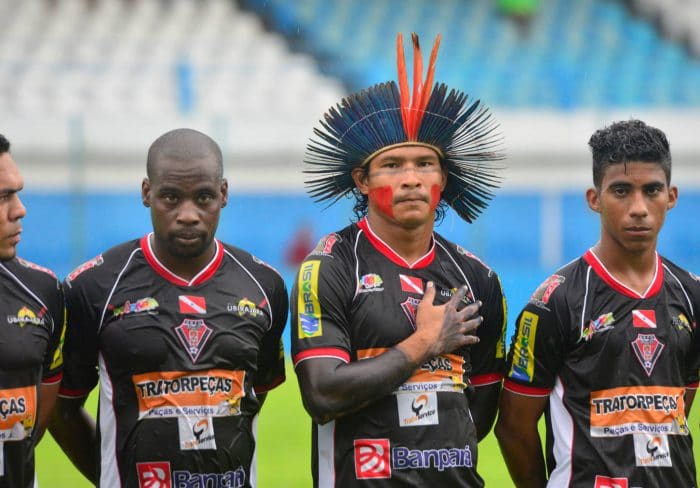 Un joueur de football autochtone en tenue de match porte une coiffe traditionnelle en plumes rouges et noires, la main sur le cœur pendant l’hymne.