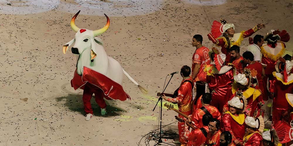 Un personnage déguisé en taureau blanc décoré de rouge danse devant un groupe de musiciens en costumes rouges ornés de dorures
