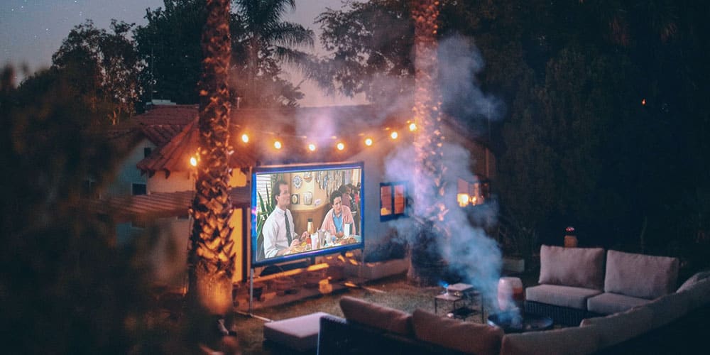 Jardin aménagé pour une projection nocturne : un écran lumineux diffuse un film tandis que des guirlandes d’ampoules suspendues entre les palmiers créent une atmosphère chaleureuse. 
