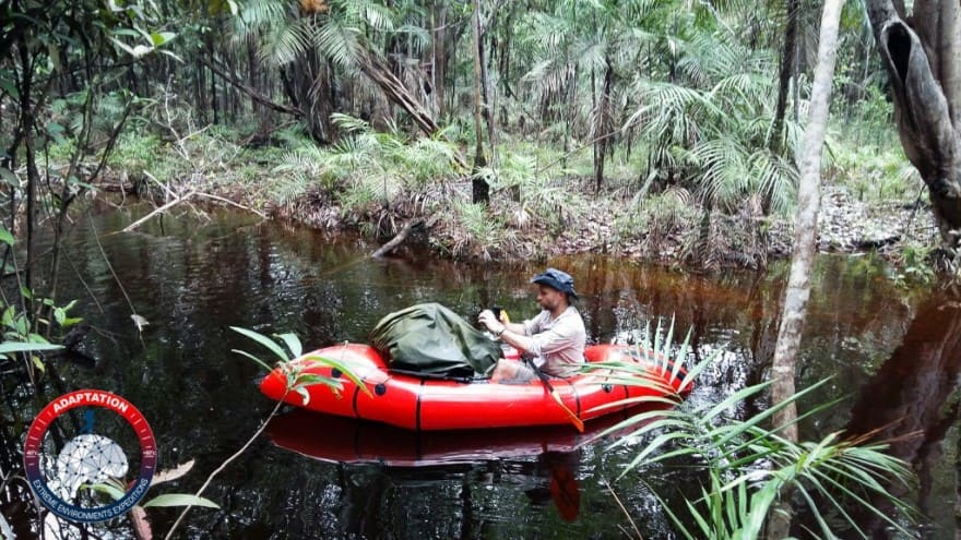 Un homme navigue sur une rivière étroite à bord d’un petit bateau gonflable rouge (packraft), en pleine forêt amazonienne.
