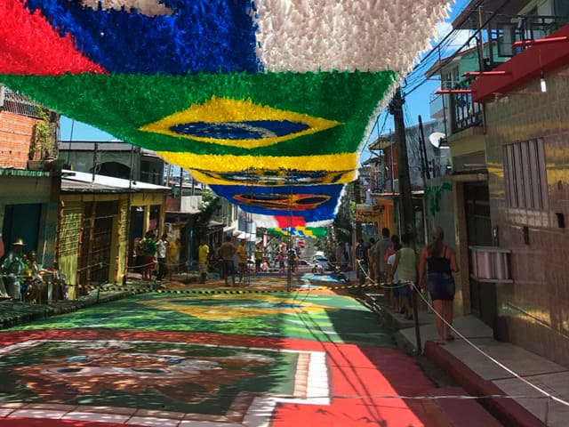 Une rue en pente est ornée de guirlandes colorées suspendues, formant le drapeau du Brésil, avec le sol également peint de motifs festifs.