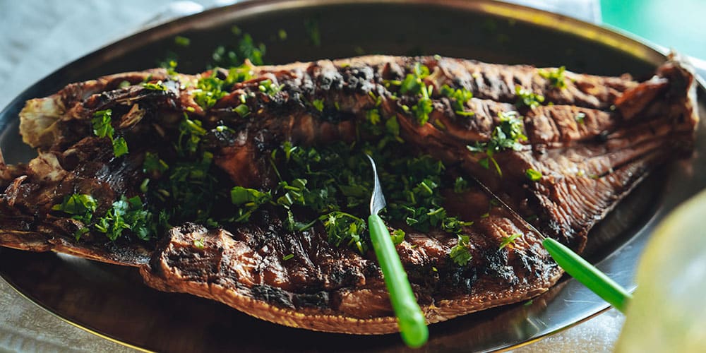 Un poisson tambaqui entier grillé, servi sur un grand plat métallique et garni de coriandre ou de persil frais.