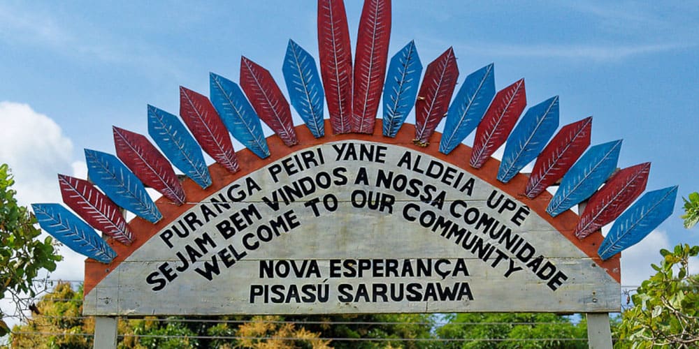 Enseigne en bois colorée de plumes rouges et bleues, surmontant l’entrée d’une communauté autochtone en Amazonie. On peut lire : Sejam bem vindos à nossa comunidade / Welcome to our community – Nova Esperança – Pisasu Sarusawa