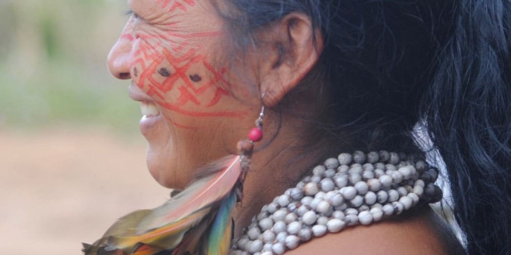 Gros plan sur le profil d’une femme autochtone souriante, portant un collier de perles naturelles blanches, une boucle d’oreille ornée de plumes colorées et des peintures traditionnelles rouges sur le visage.