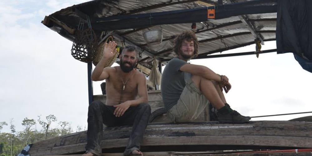 Deux hommes assis sur un radeau artisanal, l’un saluant la caméra, avec une toiture de fortune au-dessus d’eux et la forêt en arrière-plan