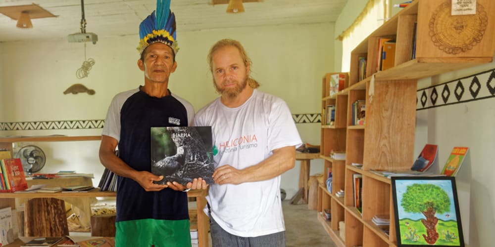 Deux hommes posent dans une bibliothèque communautaire ; l’un porte une coiffe traditionnelle à plumes bleues et jaunes, et l’autre un t-shirt “Heliconia”. Ils tiennent ensemble un livre intitulé Harpia, probablement lié à l’aigle harpie.