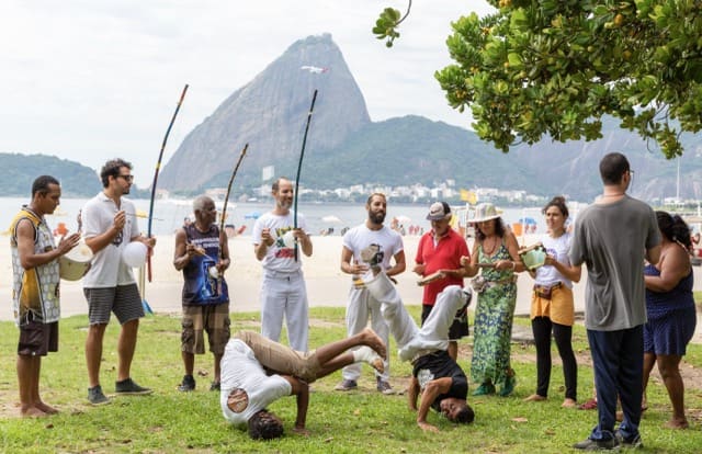 Scène de capoeira en plein air sur une pelouse en bord de mer, avec le Pain de Sucre en arrière-plan.