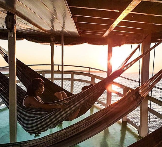 Une personne se repose dans un hamac suspendu sur le pont d’un bateau de transport fluvial, alors que le soleil se couche à l’horizon.